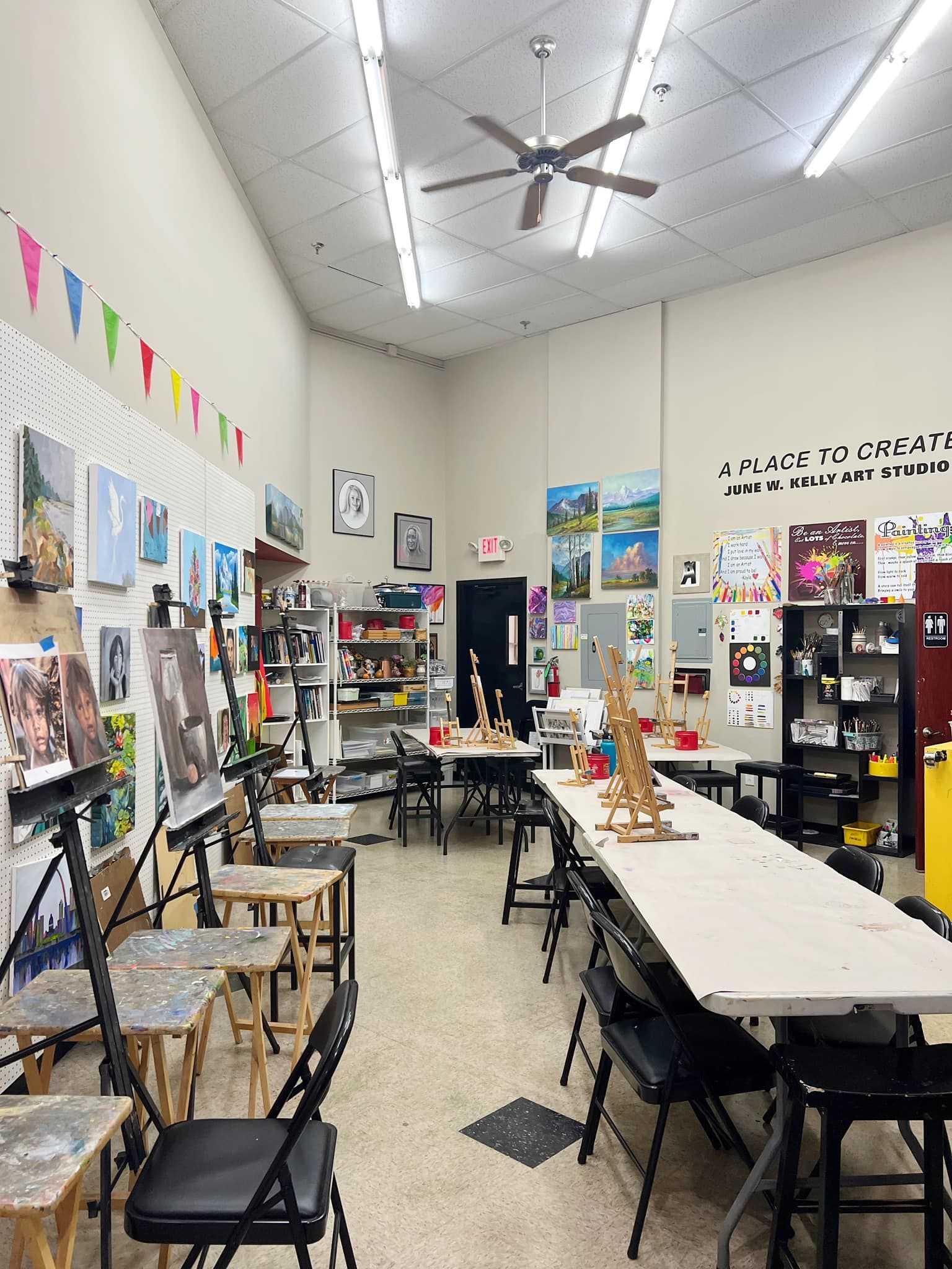 Art classroom with long tables, chairs, paintings on the walls, and shelves of supplies under a ceiling fan.