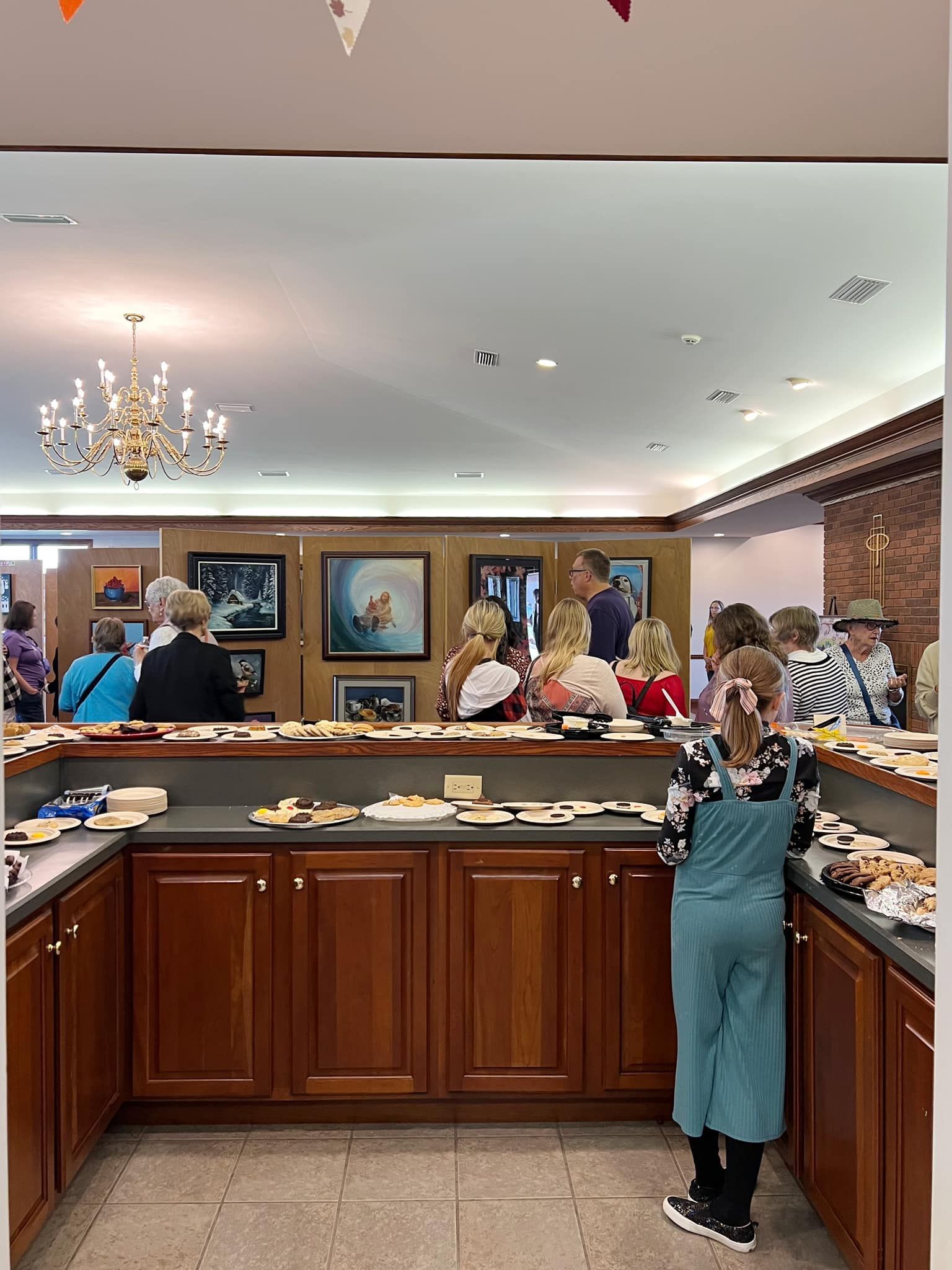 People serving pastries at a buffet counter in a bright dining room with chandeliers and framed art.