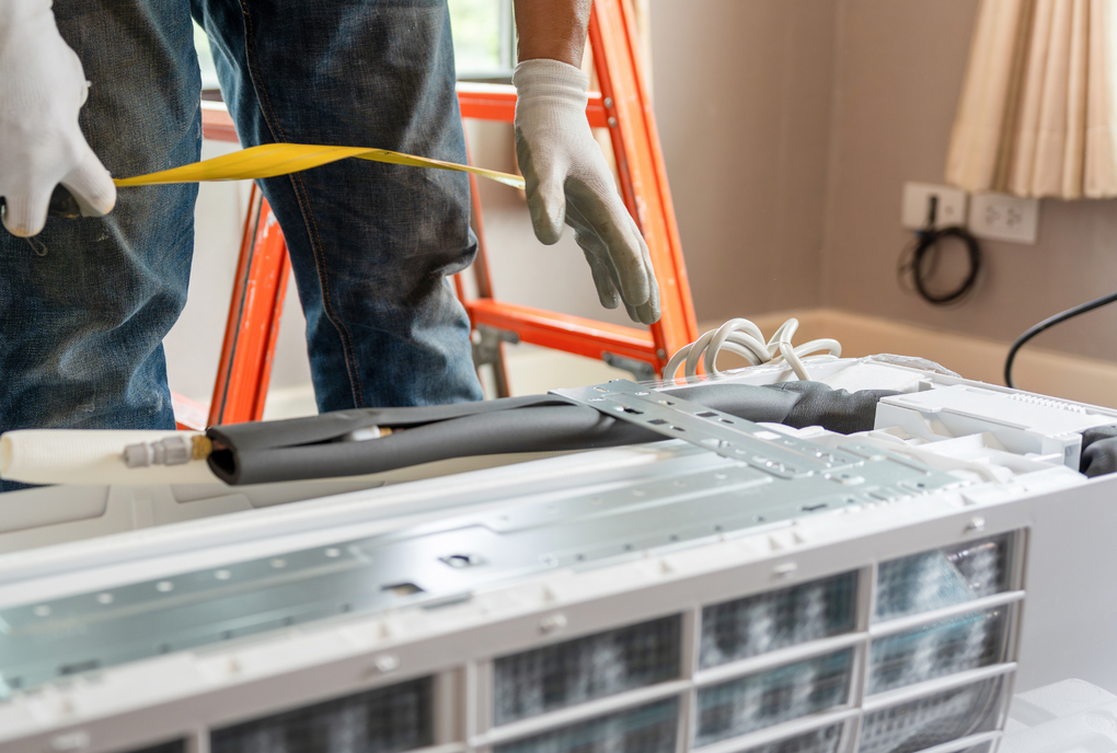 A worker in work clothes and gloves installs or repairs an indoor air conditioning unit.