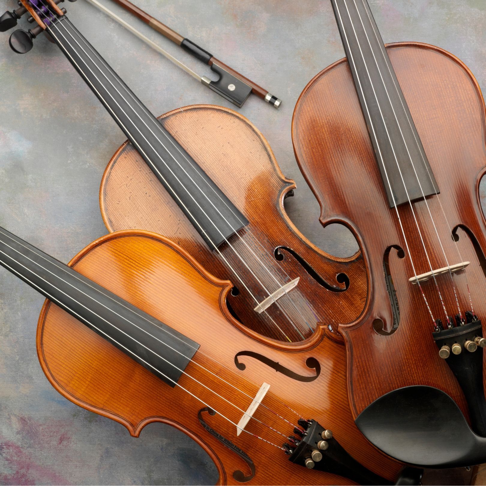 Three violins with black fingerboards and warm brown-orange bodies on a gray surface