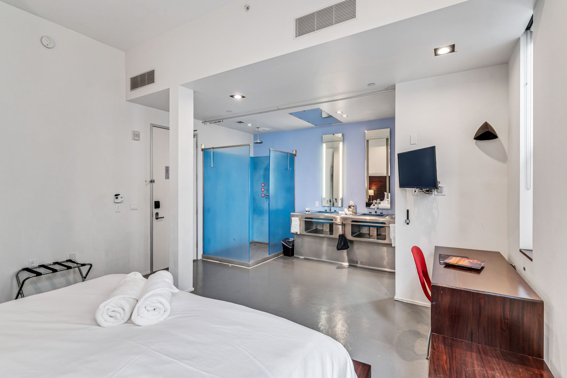 Hotel room with bed, shower, desk, and vanity. White walls and blue accents.