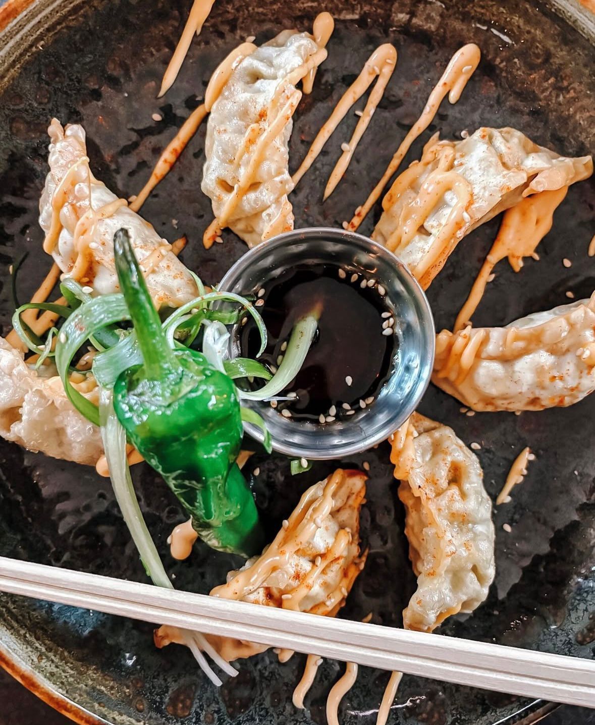 Gegrilde dumplings in een hete pan met groene saus, een dipschaaltje en metalen eetstokjes.