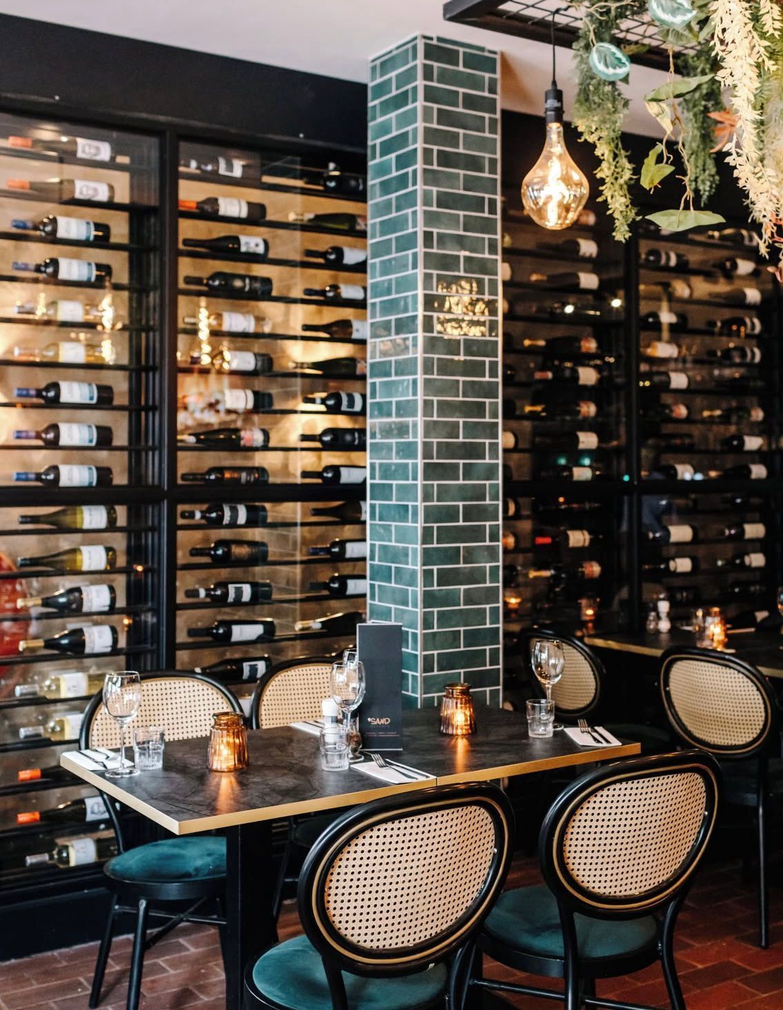 Elegant restaurantinterieur met wijnrekken, een pilaar van groene tegels en een gedekte eettafel met gevlochten stoelen.