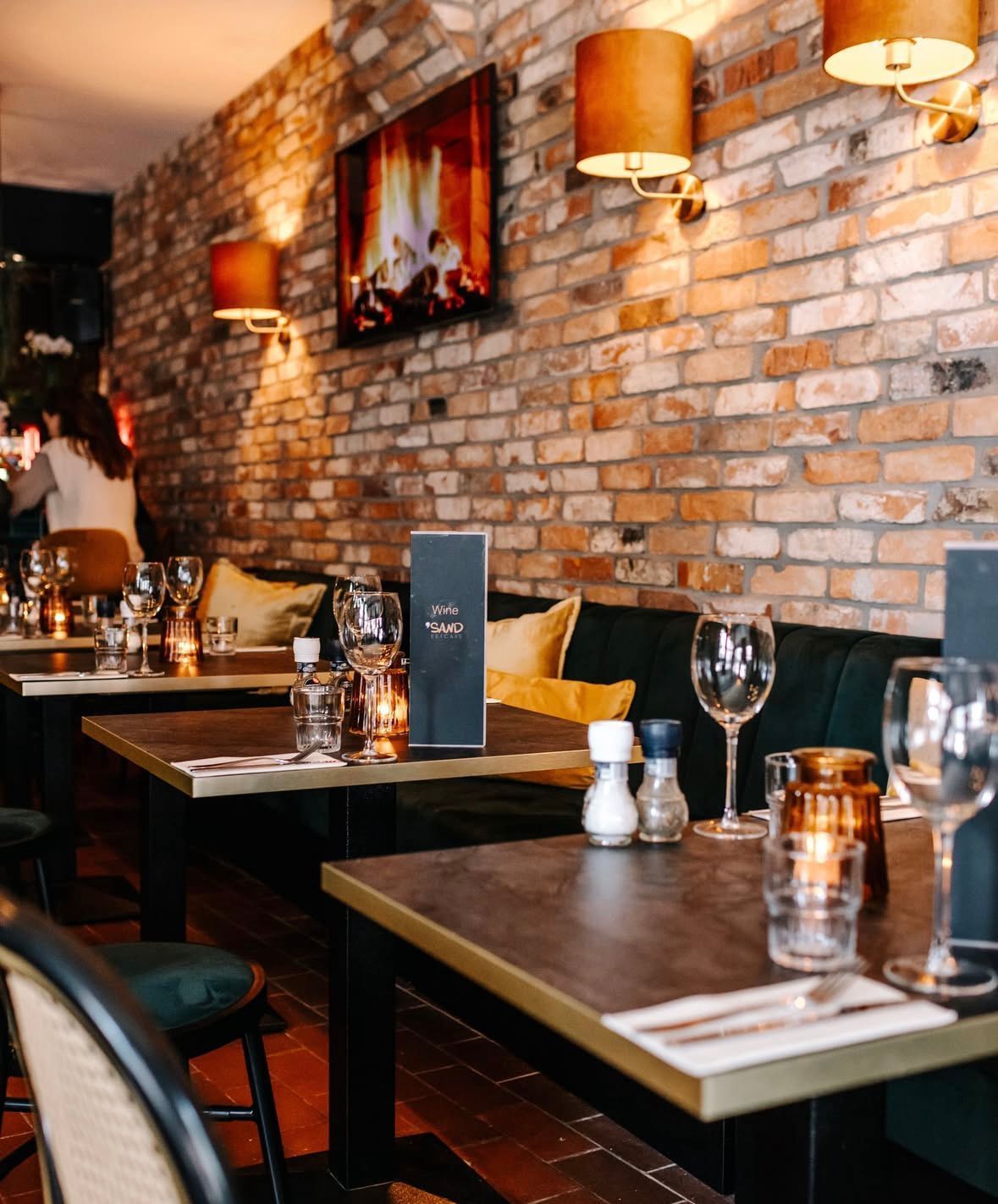 Gezellig restaurantinterieur met bakstenen muren, houten tafels, donkere banken en warme hanglampen.