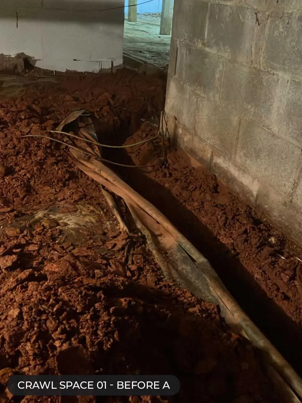 Crawl space with muddy trench and a long pipe beside a concrete foundation wall.