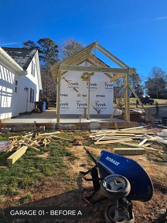 House under construction with exposed framing, scattered lumber, and a blue wheelbarrow in the yard.