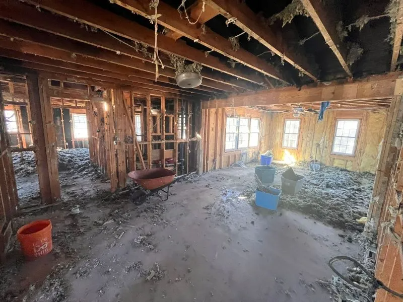 Damaged room interior with exposed ceiling joists, debris on floor, wheelbarrow, and bright windows on one wall