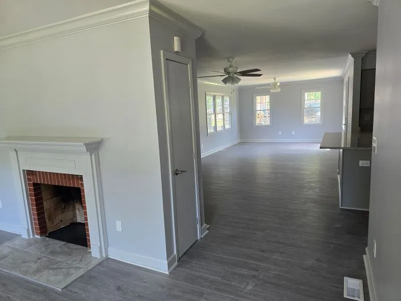 Empty living room with white fireplace, gray floors, and large windows in a bright open-plan space