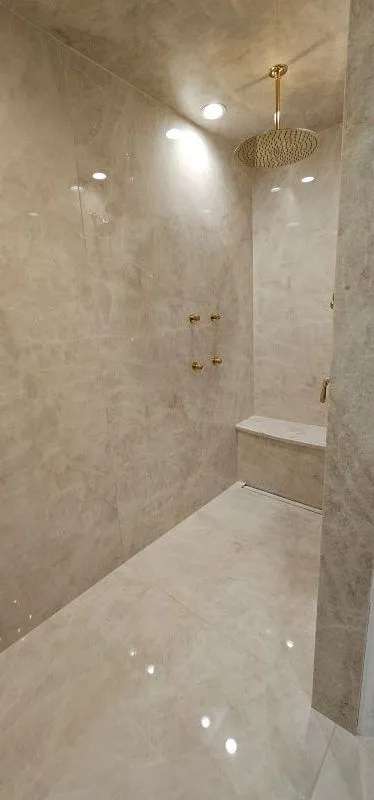 Empty marble-tiled shower with built-in bench and gold fixtures, lit by a ceiling light.