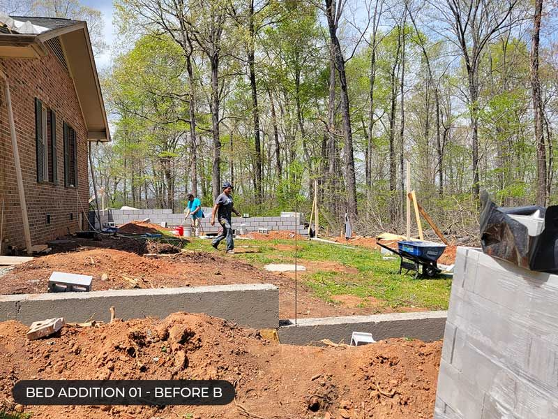 Backyard construction site with raised bed walls, dirt piles, tools, and workers near a brick house