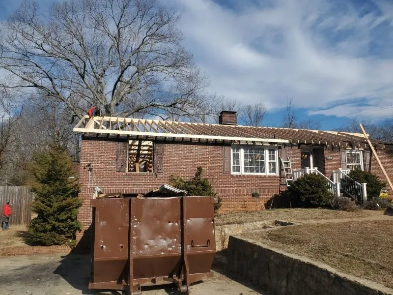 Brick house with damaged roof and a large brown dumpster in the foreground under a blue sky.