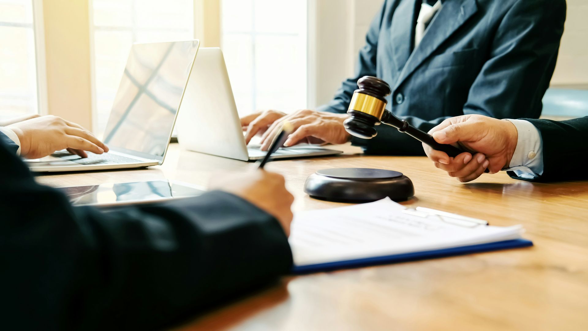 People in suits at a table, one holding a gavel, others typing on laptops and looking at documents.