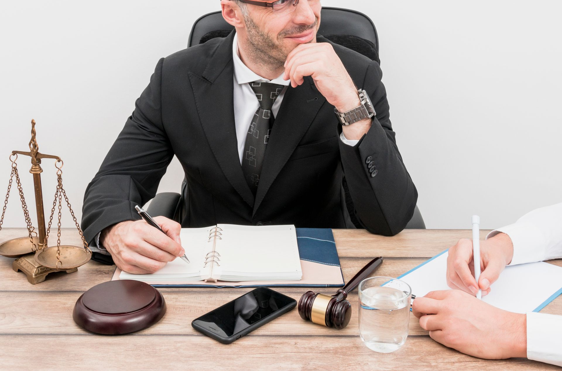 Man in suit at desk with notepad, scales of justice, and gavel; person next to him takes notes.