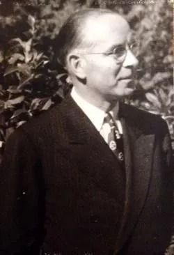 Black-and-white portrait of a man in a suit standing outdoors, facing left and wearing glasses