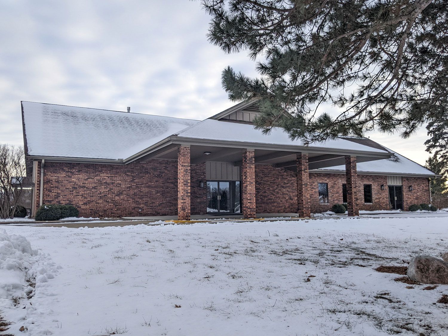 A red brick building with a covered entrance and snow-covered roof, set against a cloudy sky during winter.