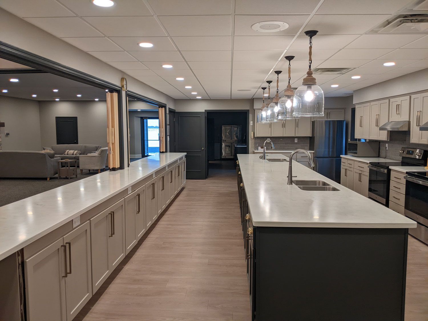 A long, modern kitchen features a spacious island with pendant lights, white countertops, and dark lower cabinets.