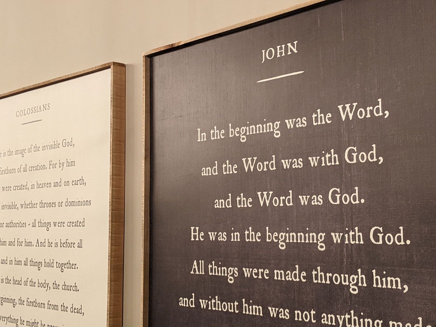 Two framed signs on a wall; one white with black text and one black with white text showing the Gospel of John, chapter 1.