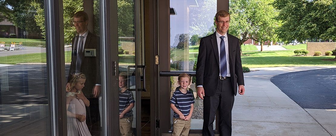 A person in a suit and children stand near glass double doors leading to a lawn.
