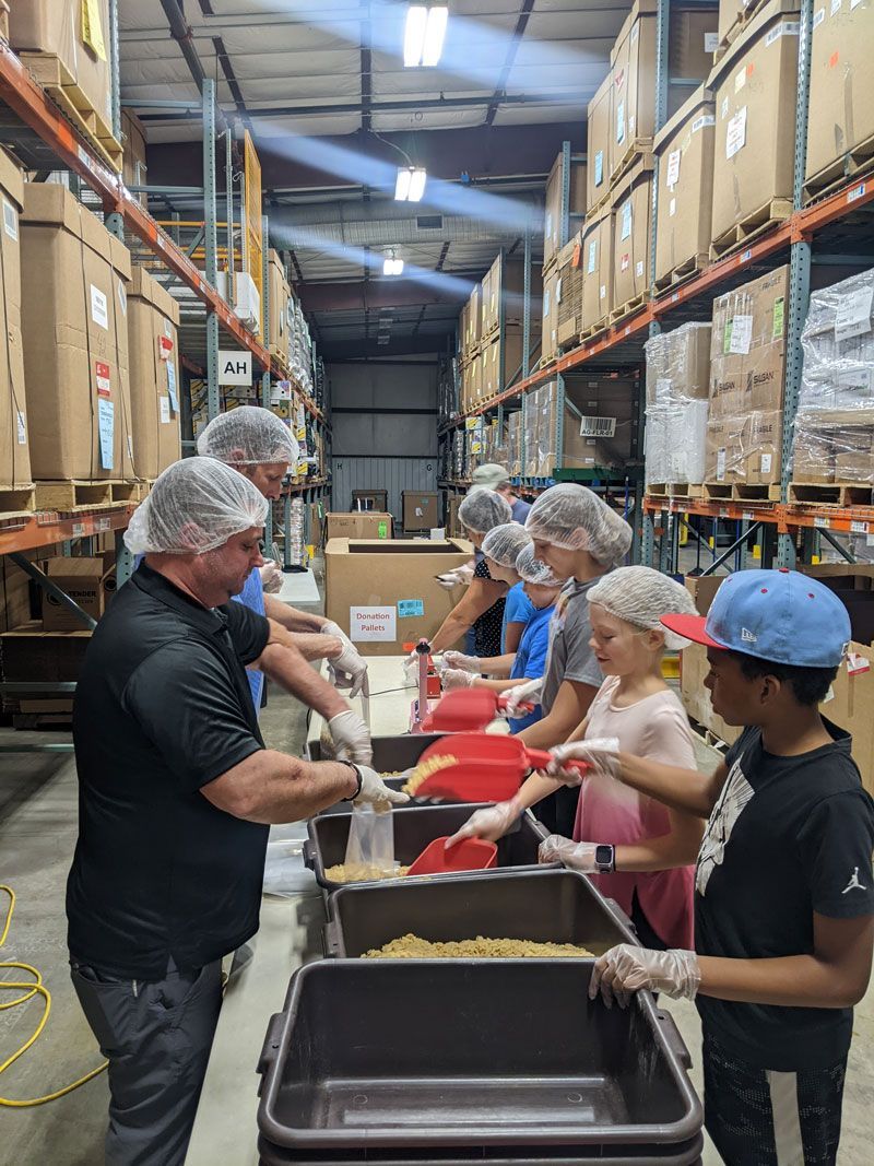 Volunteers in hairnets and gloves scoop bulk dry food into containers at a warehouse packing event.