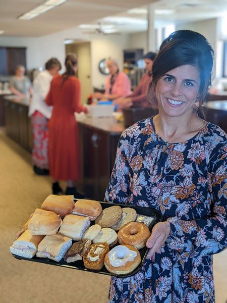 A person smiles while holding a tray of various baked goods in an office breakroom, with others standing in the background.