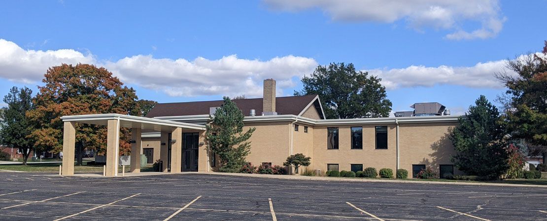 A tan, single-story brick building with a covered entrance and parking lot under a blue sky with scattered clouds.