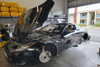 Black sports car in a garage with hood open and front wheel removed, showing repair work