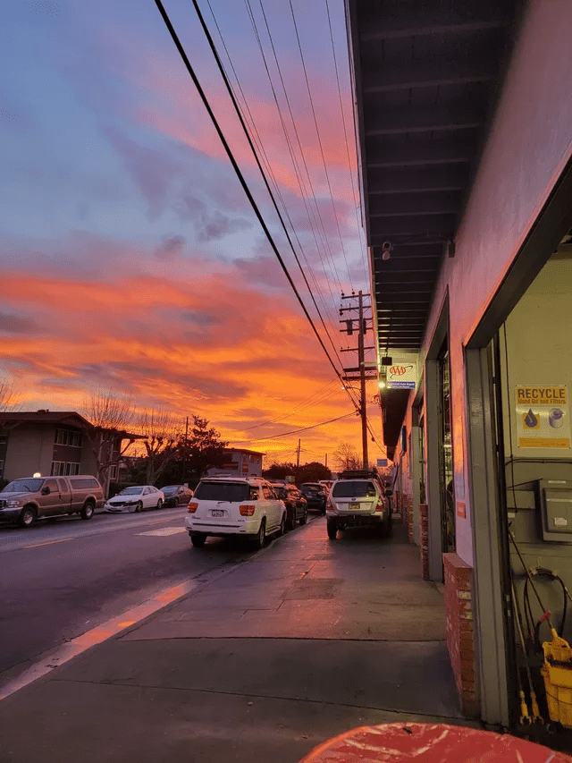 Sunset over a wet street with parked cars and shopfronts, glowing orange and pink clouds overhead
