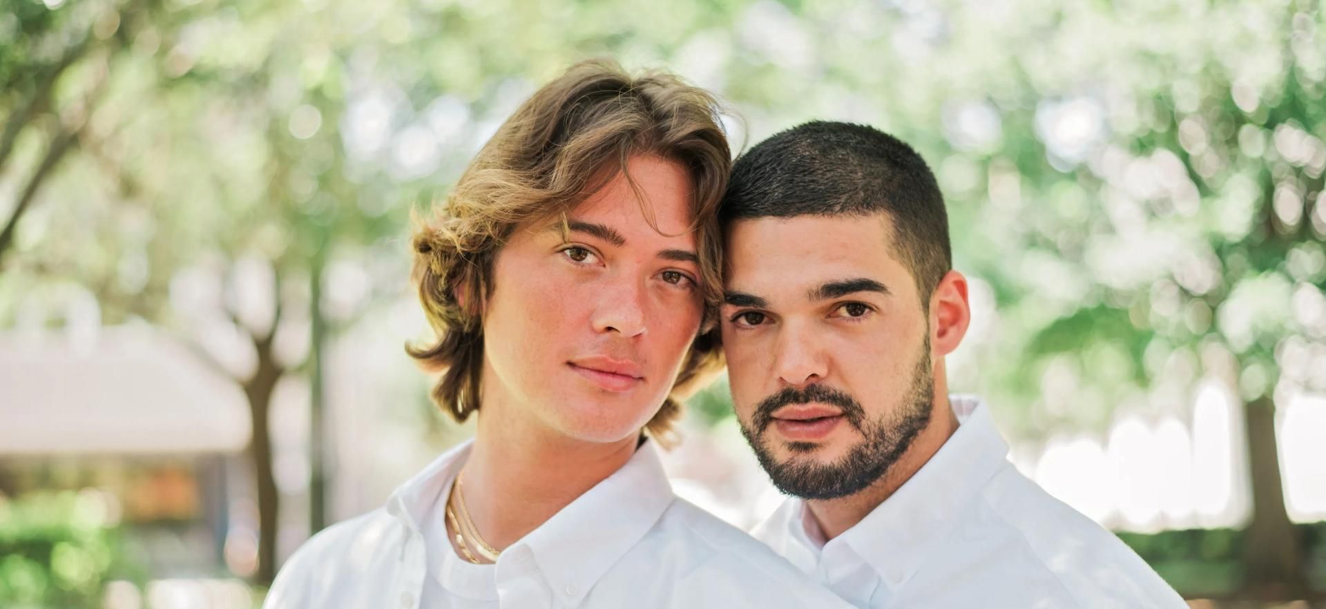 Two people in white shirts standing outdoors in a sunlit park, looking at the camera.
