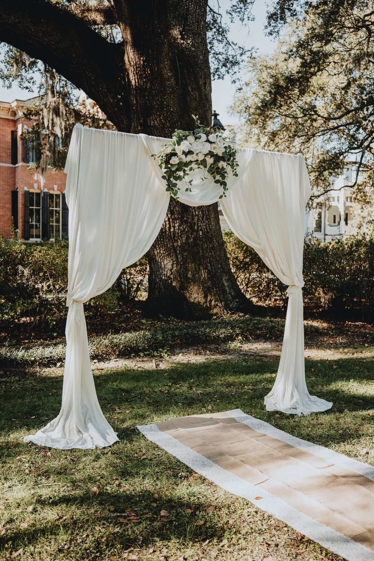 White draped wedding arch with floral centerpiece under a large tree on a grassy lawn, with an aisle runner nearby