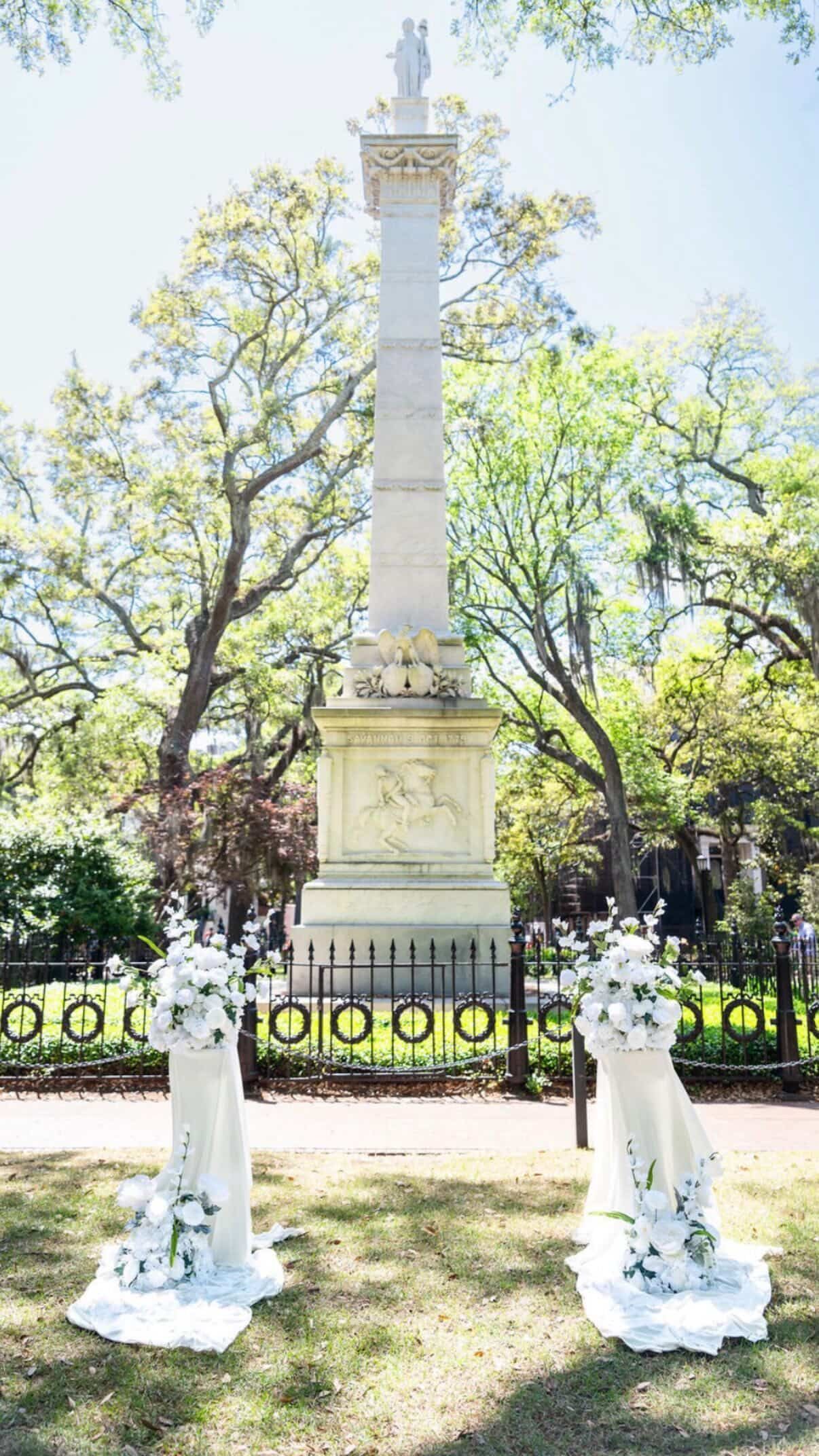 Stone memorial column in a tree-lined park, flanked by white floral arrangements and black iron fencing.