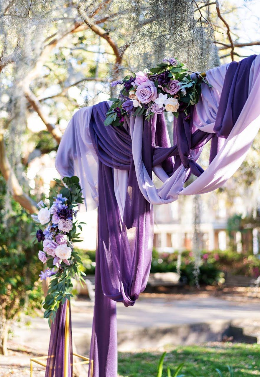 Purple and lavender fabric draped over a floral outdoor arch in a wooded setting
