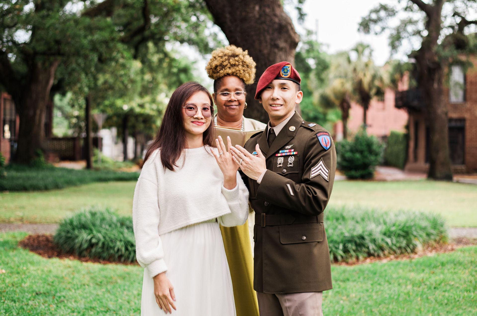 Three people posing outdoors in a garden, one in a white dress, one in a yellow dress, and one in military uniform.