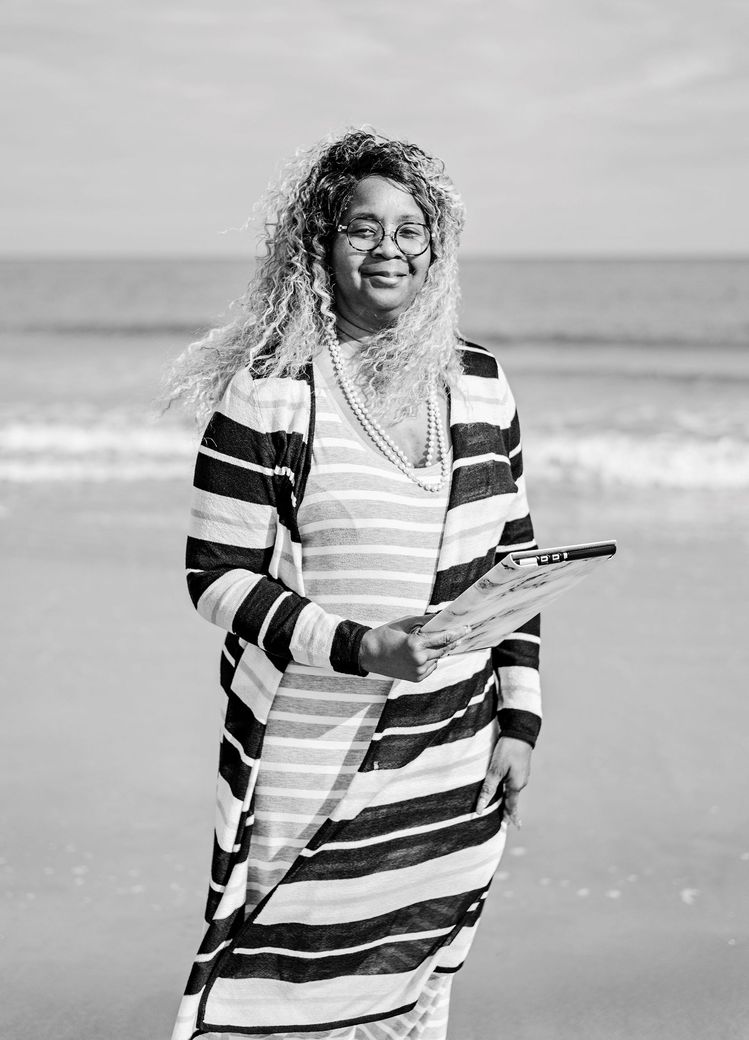 Person standing on a beach holding a book, wearing a striped outfit and smiling.