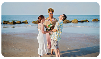 Three people in formal wear pose on a beach, holding flowers with the ocean behind them.