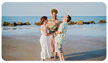 Three people in formal wear pose on a beach, holding flowers with the ocean behind them.