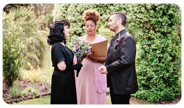Couple exchanging vows with officiant in a garden, holding a bouquet and clipboard.