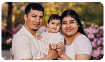 Smiling family of three sitting outdoors, with pink flowers in the blurred background.
