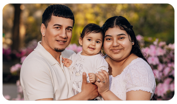 Smiling family of three sitting outdoors, with pink flowers in the blurred background.