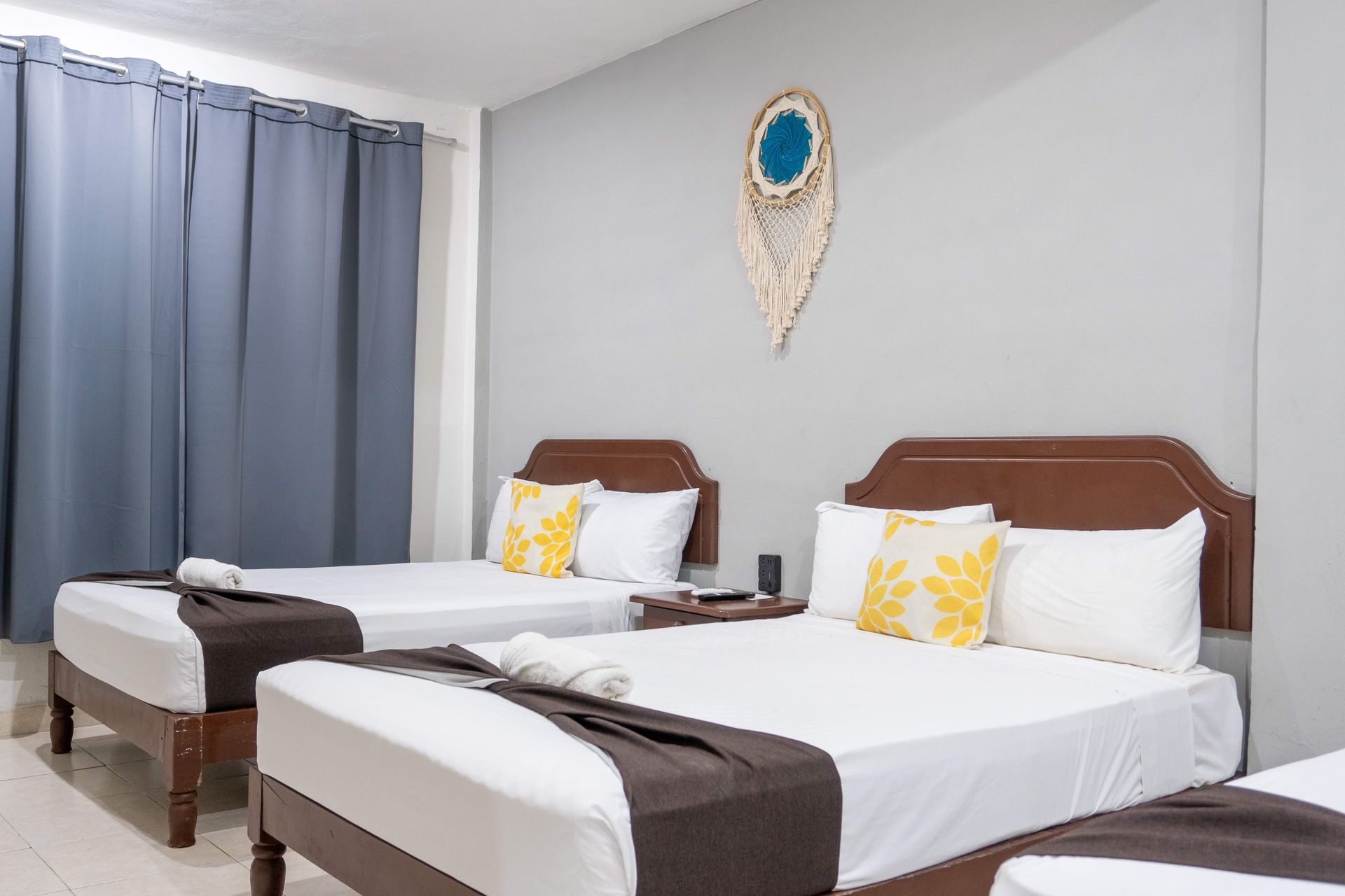 Habitación luminosa con dos camas individuales, sábanas blancas, cabeceros marrones y cortinas azules.