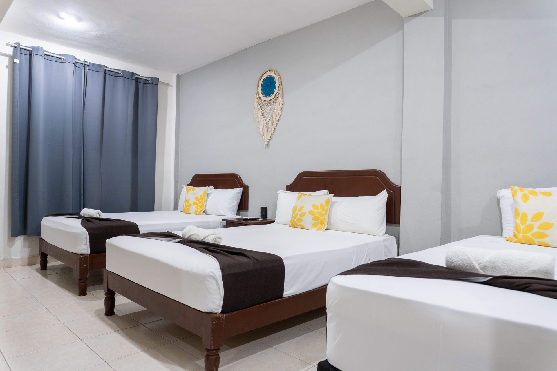 Habitación de hotel luminosa con cuatro camas individuales bien hechas, ropa de cama blanca y marrón y cortinas azules.
