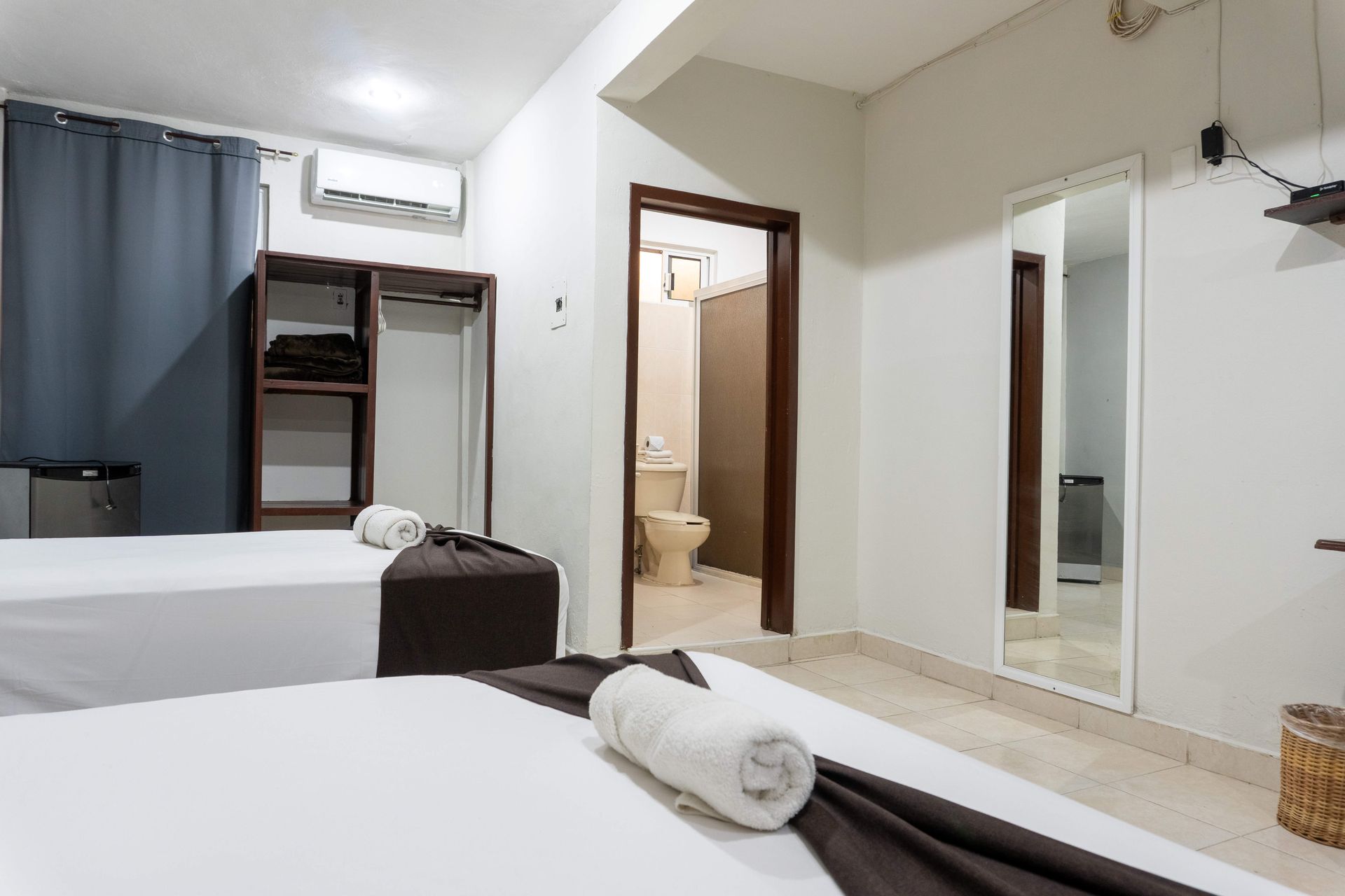 Habitación luminosa con dos camas individuales, puerta de baño, espejo y ventana con cortinas.