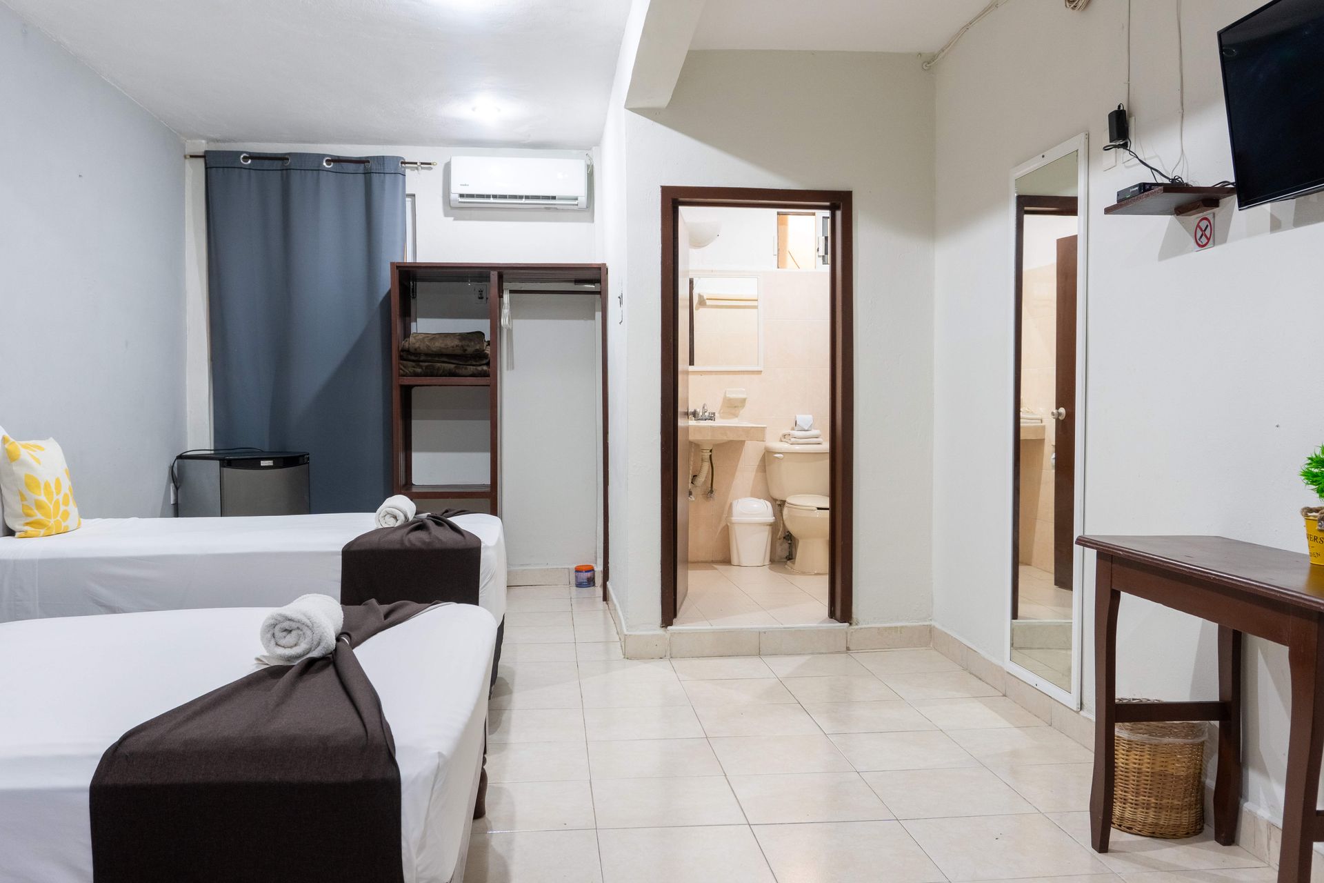 Habitación con dos camas individuales, suelo de baldosas, baño abierto y escritorio en espacio luminoso y moderno.