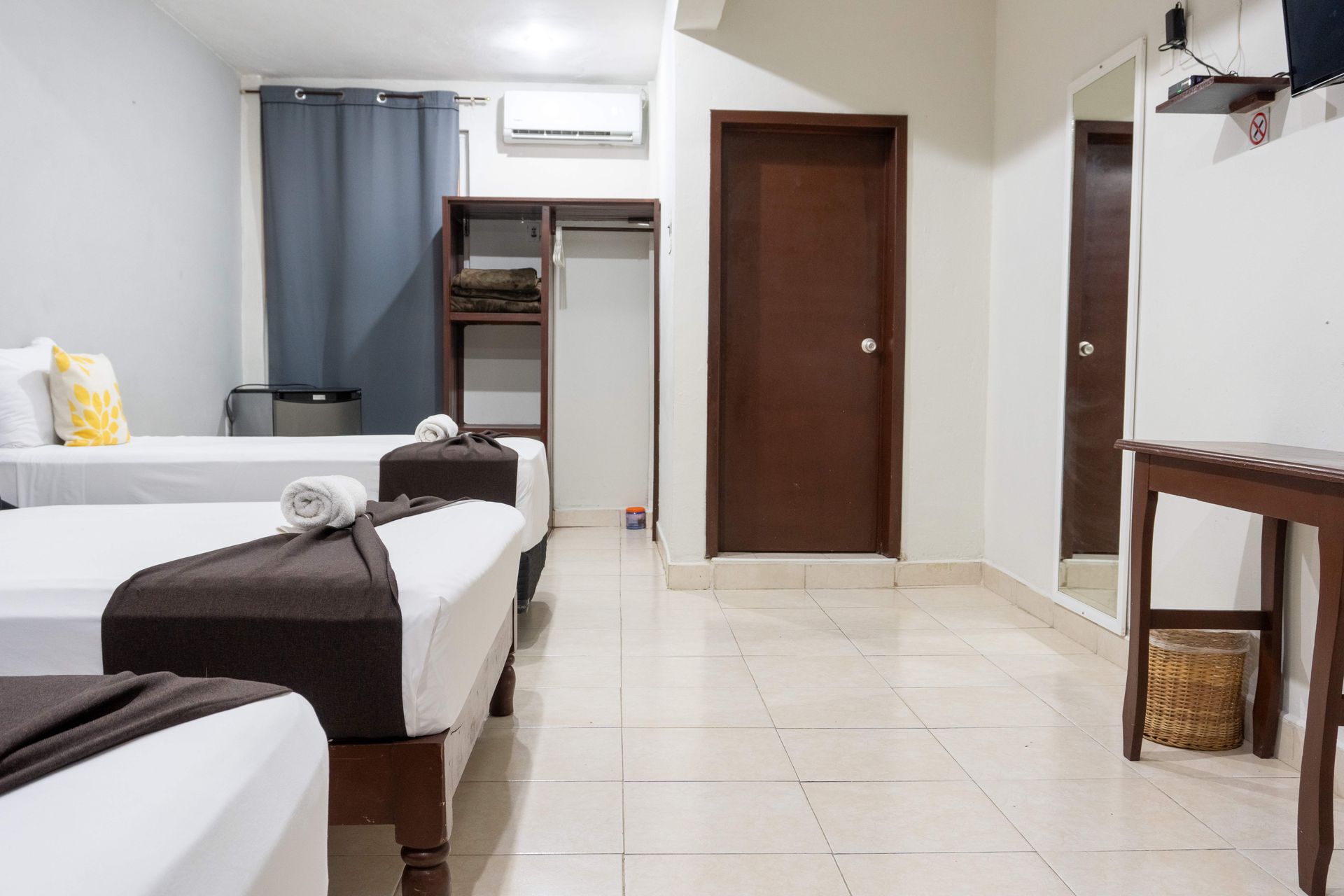 Habitación luminosa con dos camas, un armario, puerta marrón, aire acondicionado y una mesita con espejo.