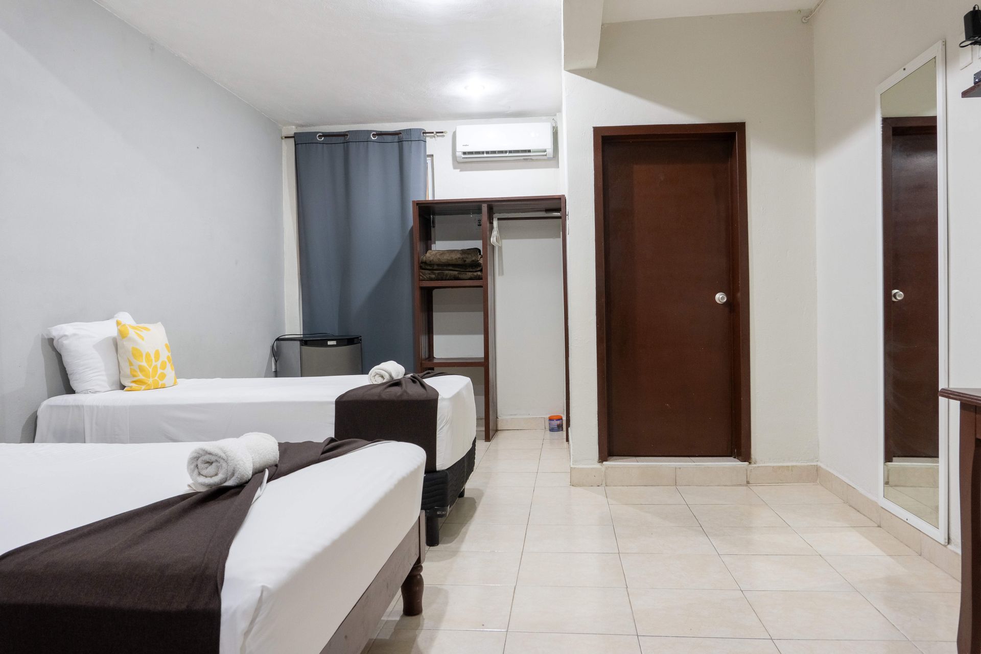 Habitación pequeña tipo hotel con dos camas, suelo de baldosas, cortinas, aire acondicionado y puerta abierta.