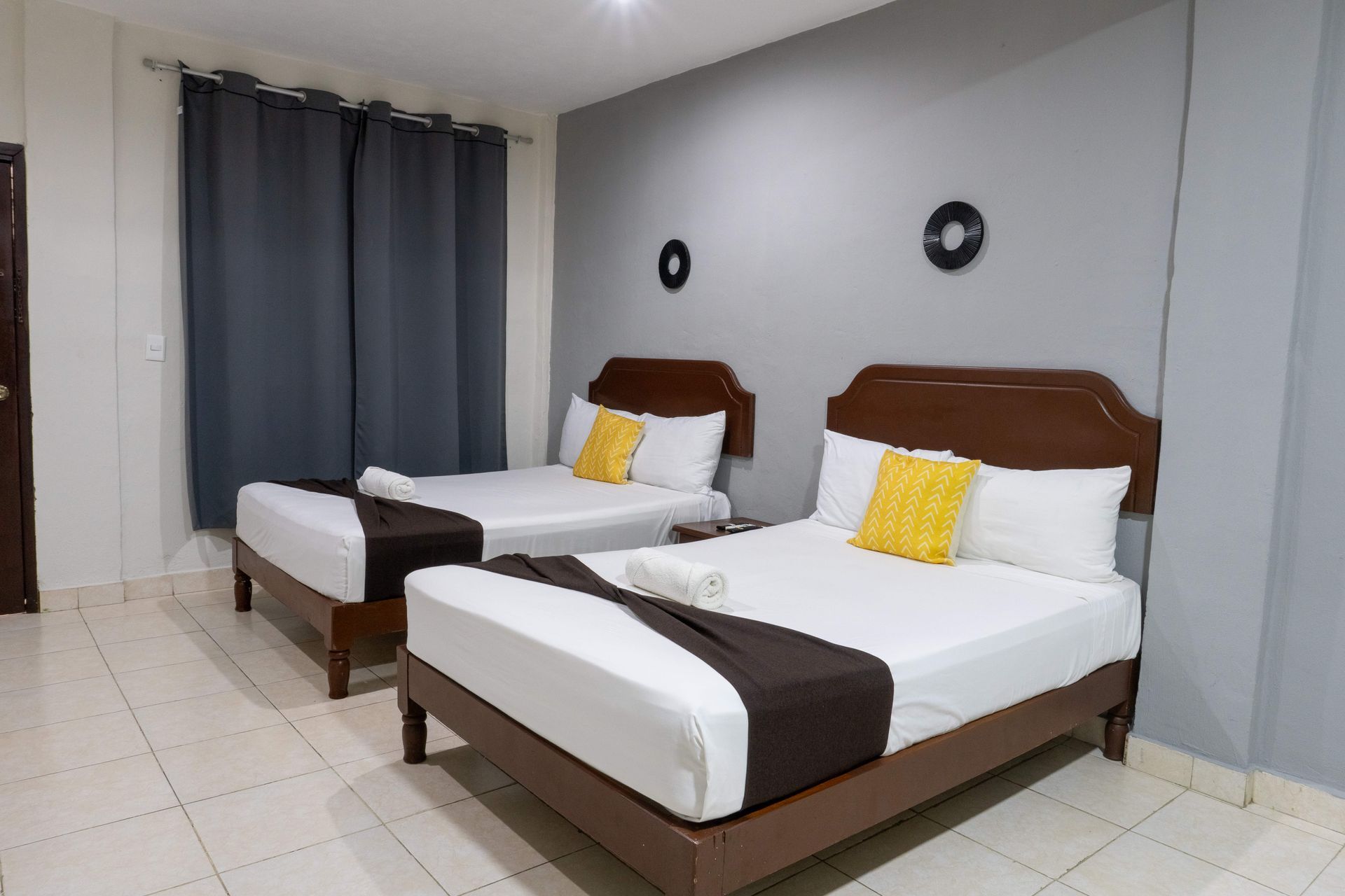 Habitación minimalista con dos camas individuales, paredes grises, suelo de baldosas blancas y cortinas oscuras.