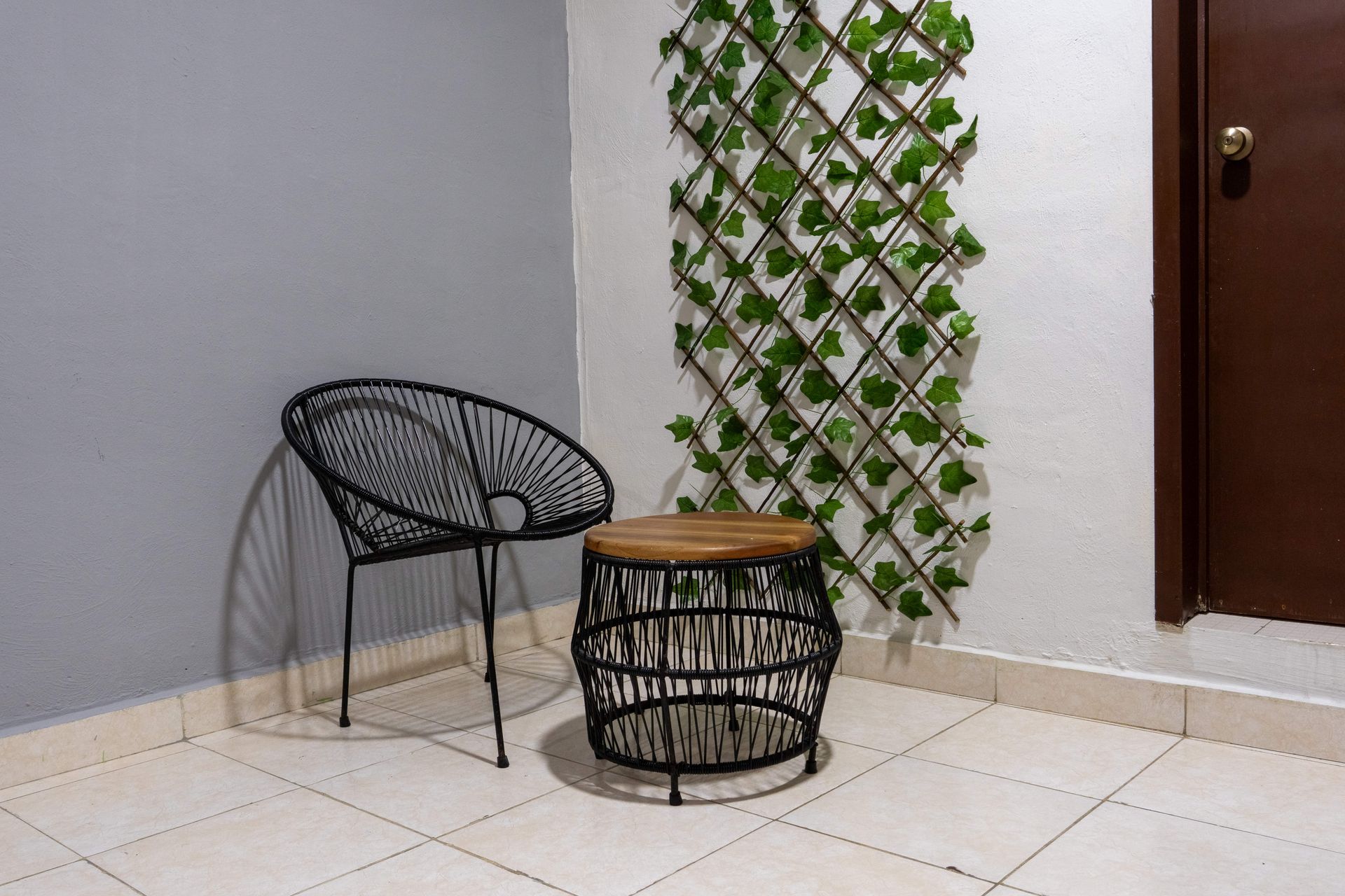 Patio de esquina con silla de alambre negro, taburete tejido y celosía con plantas trepadoras sobre suelo de baldosas.
