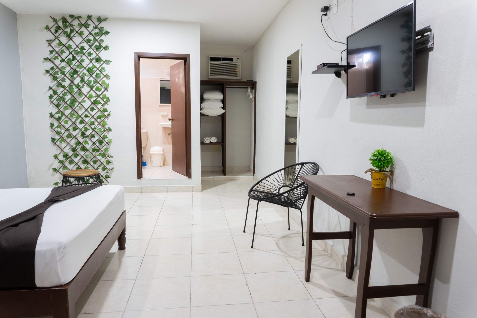 Habitación de hotel pequeña con cama, escritorio, televisor montado en la pared, silla, armario y puerta de baño.