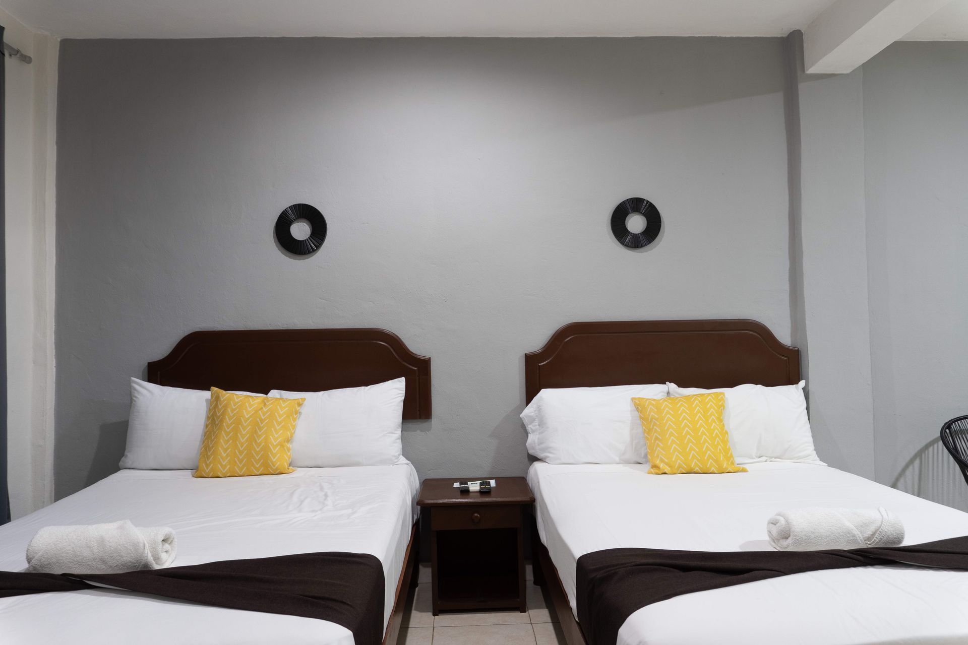 Habitación de hotel con dos camas individuales, sábanas blancas, almohadas amarillas y una pared gris con luces redondas.