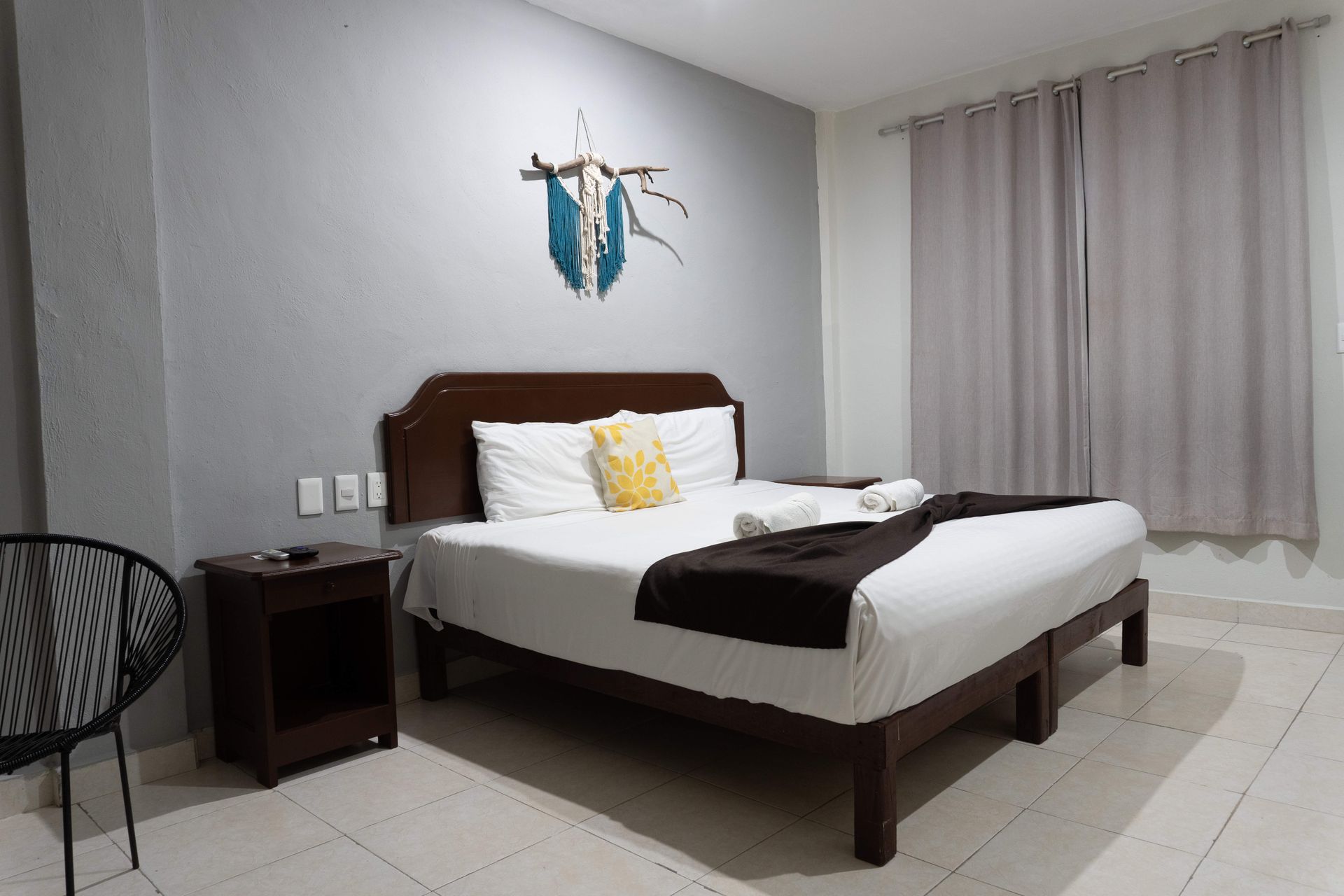 Habitación ordenada con cama, cuadro en la pared, silla negra, mesita de noche y cortinas grises.