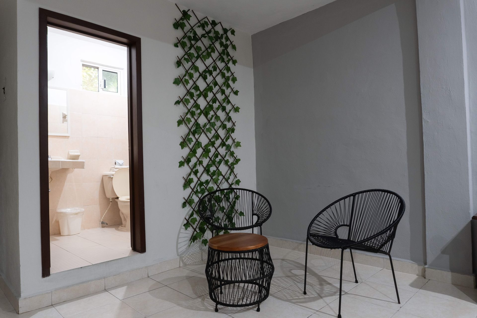 Patio minimalista con dos sillas negras, una mesa pequeña y una pared cubierta de enredaderas junto a una puerta.